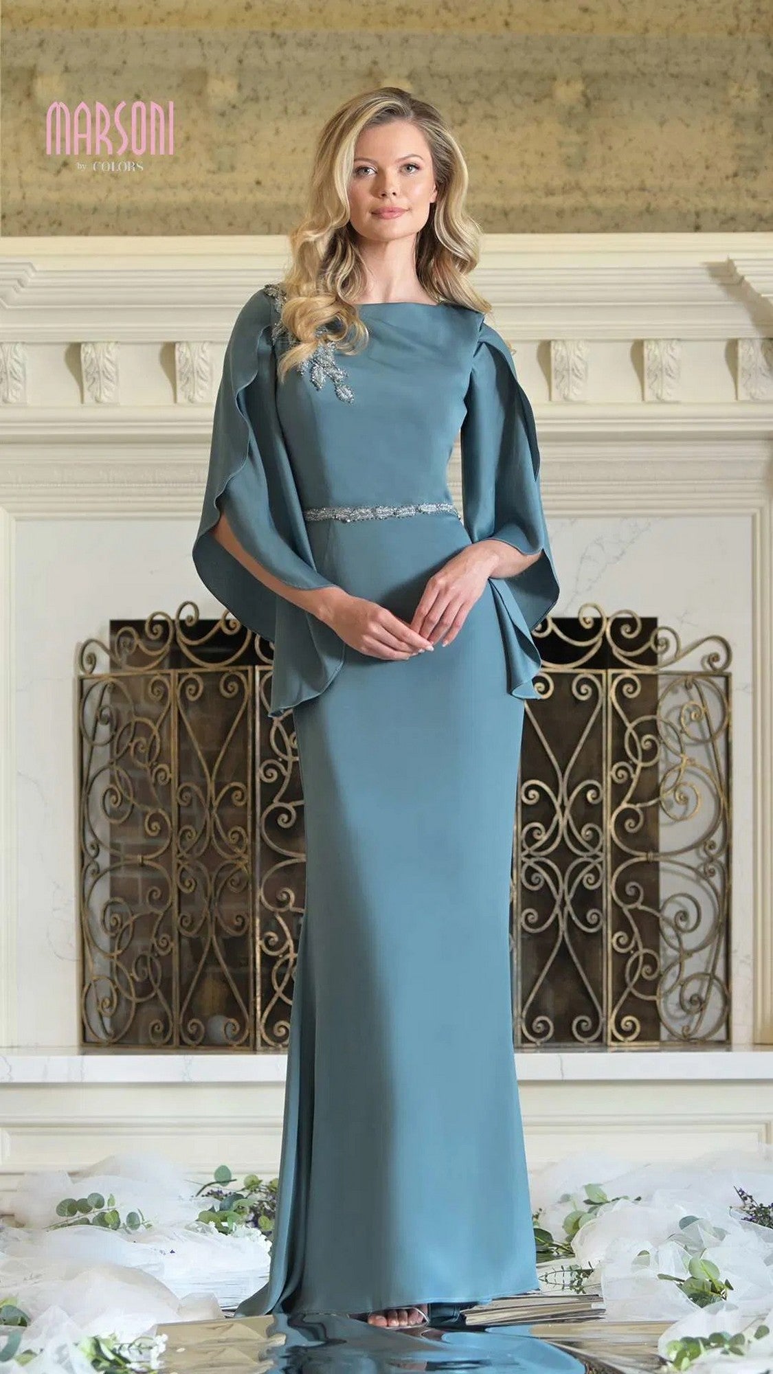 A woman stands indoors wearing the Marsoni MV1348 long formal dress—a floor-length blue gown featuring cape sleeves and beaded appliques at the waist and shoulder.